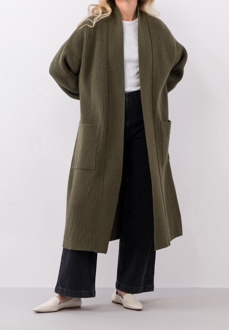 Oliwkowy żakiet ze żłobieniami, z długim, otwartym przodem, szerokimi rękawami i dwiema przednimi kieszeniami. Połączony z czarnymi spodniami w szerokim kroju i beżowymi butami.