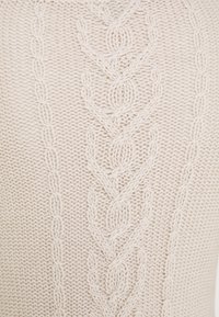 Beige stickad tröja med ett centralt flätat mönster med sammanflätade öglor och texturerade stygn, som visar en mjuk, chunky kvalitet.