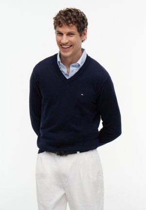 Homme aux cheveux bouclés souriant, portant un pull bleu marine à col en V sur une chemise bleu clair boutonnée et un pantalon blanc, sur fond blanc.