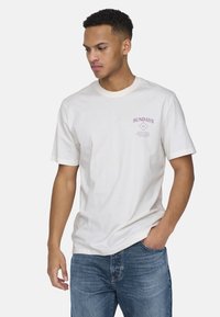 White cotton t-shirt with short sleeves, featuring printed maroon text "SUNDAYS" and a geometric design on the front. Fitted silhouette.