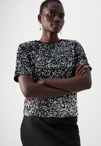 Haut à manches courtes en sequins noirs avec un col rond, présentant une surface texturée et orné de sequins argentés brillants.