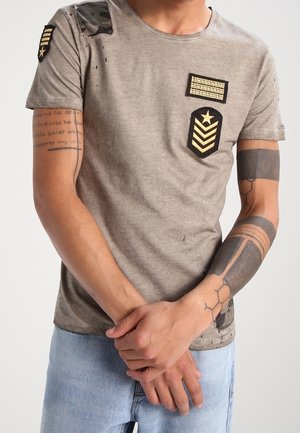 Homme portant un t-shirt marron clair usé avec des écussons de style militaire et un jean bleu, bras croisés montrant des tatouages géométriques et en écriture.