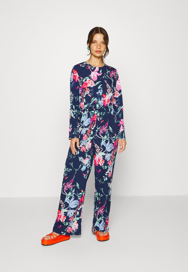 Conjunto floral de duas peças com mangas longas e calças largas, num fundo azul-marinho, decorado com flores vibrantes em rosa, roxo e verde. Calçado laranja.