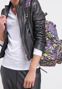 Osoba oblečená v černé kožené bundě, bílé dlouhé tričko a nese barevný batoh s vzorem přes jedno rameno.