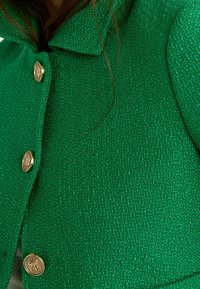 Giacca verde strutturata con colletto e bottoni dorati, con un motivo intrecciato e una vista ravvicinata del suo materiale.