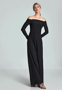 Schwarzer Jumpsuit mit schulterfreiem Schnitt aus dehnbarem Stoff, langen Ärmeln und fließender Silhouette, kombiniert mit schwarzen Riemchensandalen.