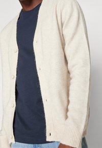 Beige cardigan i mjukt material, med sex knappar och ribbstickade muddar, lagd över en marinblå t-shirt. Avslappnad, öppen design.