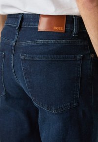 Σκούρο μπλε τζιν παντελόνι με πίσω τσέπη και καφέ δερμάτινο πατινί που φέρει την ετικέτα BOSS, σε ένα άτομο που φοράει λευκό πουκάμισο.