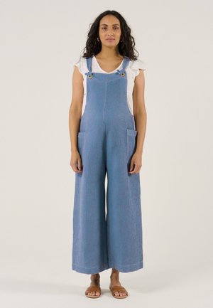 Femme aux longs cheveux foncés portant une chemise blanche à manches courtes sous une salopette large bleue texturée et des sandales plates marron, debout.