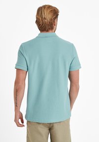 Polo shirt in light blue, textured fabric, short sleeves, and a subtle collar. Visible tattoo on the left arm. Worn with light-colored shorts.