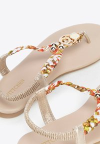 Gouden sandalen hebben gevlochten banden in oranje, groen en wit. Glad beige voetbed met decoratieve accenten en metalen hardware.