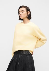 Jeune personne aux cheveux courts et foncés portant un pull ample jaune pâle et une jupe plissée noire taille haute avec ceinture, se tenant de profil.