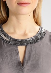 Close-up van een persoon die een grijze top draagt met een bewerkte halslijn en een V-vormige uitsnijding aan de voorkant.