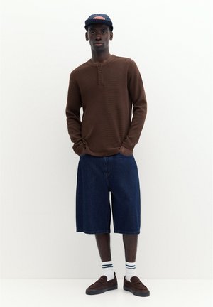 Un jeune homme porte une chemise marron à manches longues, un short en jean bleu foncé, des chaussettes blanches à rayures, des mocassins foncés et une casquette bleu marine, debout sur un fond blanc.