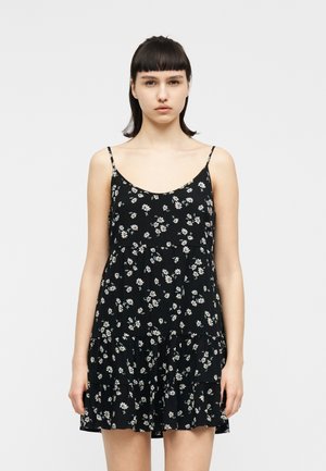 Tânără cu păr scurt și închis la culoare, purtând o rochie neagră fără mâneci cu imprimeu floral de margarete albe, stând în picioare pe un fundal simplu, deschis la culoare.