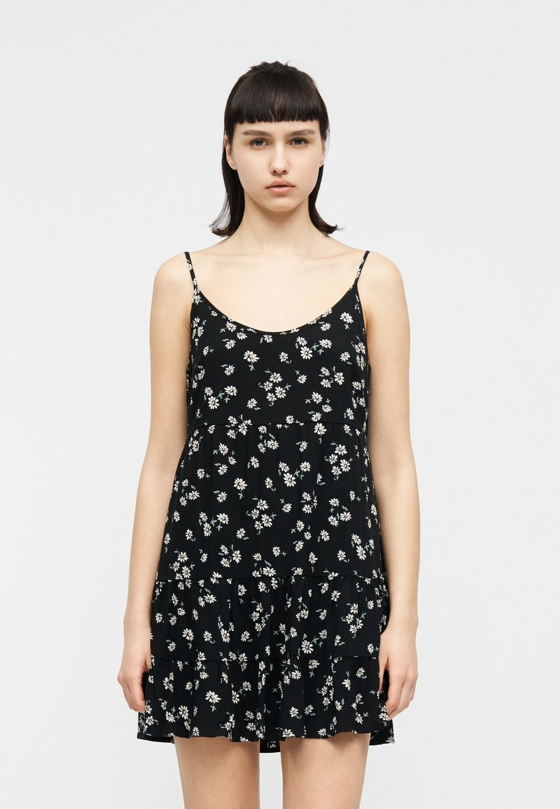 Νεαρή γυναίκα με κοντά σκούρα μαλλιά, ντυμένη με αμάνικο μαύρο φόρεμα με λευκό σχέδιο λουλουδιών μαργαρίτας, στέκεται μπροστά σε απλό ανοιχτόχρωμο φόντο.