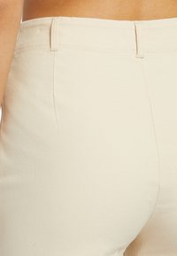 Nahaufnahme einer Person, die hochgeschnittene, cremefarbene Hose mit Gürtelschlaufen und einer sichtbaren Naht am Bund trägt.