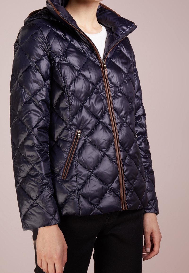Marinblå vadderad pufferjacka med en glänsande yta, huva och bruna dragkedjedetaljer. Har sidofickor med dragkedja och en åtsittande design.