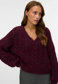 Pull en mailles torsadées bordeaux avec un décolleté en V, des manches surdimensionnées et un ourlet côtelé. Texture douce avec des motifs de tricot prononcés.