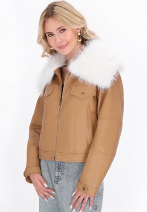 Donna bionda che indossa una giacca di pelle beige con un ampio colletto di pelliccia bianca, jeans chiari e orecchini d'oro, mentre sorride dolcemente verso la camera.