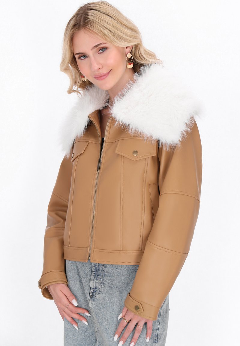 Donna bionda che indossa una giacca di pelle beige con un ampio colletto di pelliccia bianca, jeans chiari e orecchini d'oro, mentre sorride dolcemente verso la camera.