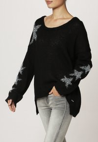 Pull en tricot noir avec design d'étoile argentée sur les manches, col rond et manches longues oversized. Présente des fentes sur les côtés et une coupe décontractée.