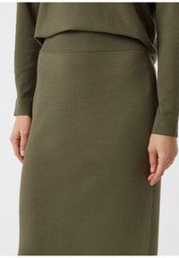 Oliwkowo zielona dzianinowa spódnica z prążkowanym paskiem w talii i gładką fakturą, w zestawie z dopasowanym topem z długim rękawem. Zbliżenie na dłonie na talii.