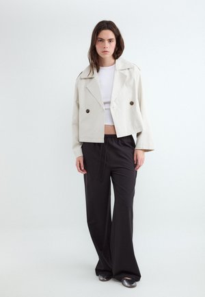 Femme debout portant une veste beige cropped à double boutonnage, un crop top blanc, un pantalon noir ample et des chaussures plates argentées sur fond uni.