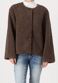 Person klädd i en brun strukturerad fleecejacka med vida ärmar över ljusblå jeans och en vit polotröja.