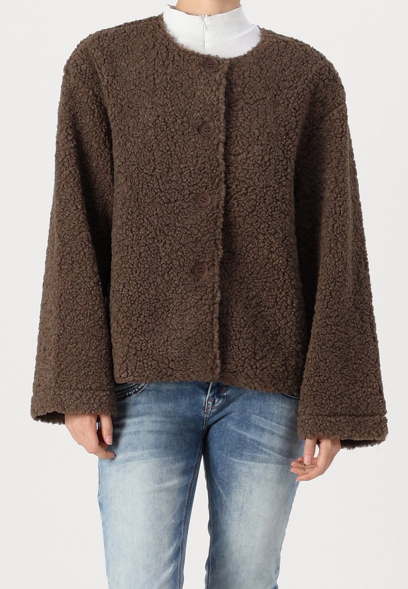 Person klädd i en brun strukturerad fleecejacka med vida ärmar över ljusblå jeans och en vit polotröja.