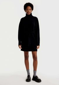 Schwarzes Strickkleid mit Rollkragen und tailliertem Gürtel. Hat lange Ärmel und einen gerippten Saum. Getragen mit schwarzen Plateauschuhen und grauen Socken.