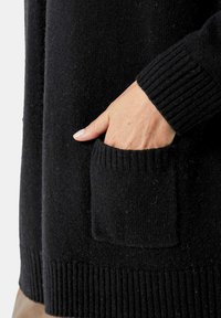 Schwarzer Strickpullover mit gerippten Bündchen und einer aufgesetzten Tasche. Der Stoff hat eine weiche Textur und sorgt für ein gemütliches, warmes Aussehen.