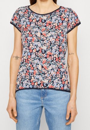 Vrouw met een korte mouw marineblauwe top met een dicht bloemenpatroon van rode, roze en lichtblauwe bloemen, gecombineerd met lichtblauwe spijkerbroek.