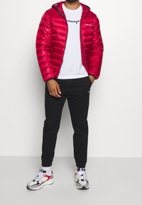 Rød pufferjakke med hætte, med lynlås lukning og skinnende overflade. Bæres over en hvid t-shirt og sorte joggers, parret med sneakers.