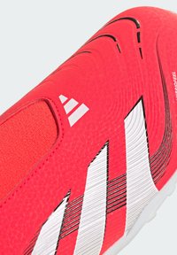 Zapato de fútbol rojo brillante con parte superior sintética texturizada, franjas blancas, cuello elástico tipo calcetín y líneas de acento negras. Se destaca la tecnología HybridFeel.