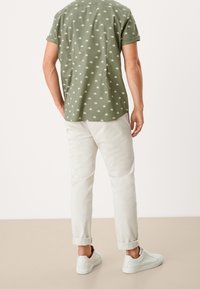 Camisa verde de manga corta con estampado de palmeras blancas en la parte posterior, combinada con pantalones color crema arremangados y zapatillas blancas.