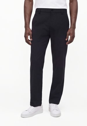 Homme portant un pantalon noir à jambes droites et des baskets blanches, debout devant un fond blanc uni.