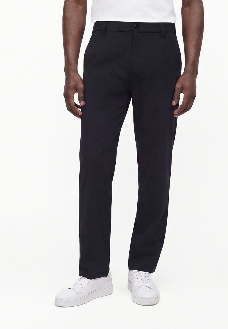 Homme portant un pantalon noir à jambes droites et des baskets blanches, debout devant un fond blanc uni.