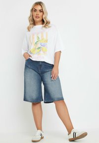 Valkoinen t-paita, jossa on sitruuna graafinen kuva, yhdistettynä sinisiin farkku Bermuda-shortsit. Valkoiset lenkkarit mustilla yksityiskohdilla täydentävät asua.