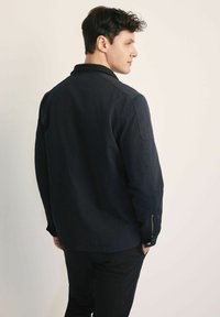 Granatowa kurtka z lekkiego materiału, z długimi rękawami, płaskim kołnierzem i zapinanymi mankietami, pokazana od tyłu.