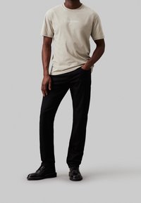 Beige bomulds t-shirt med "Calvin Klein" logo, parret med sorte bukser og sorte lædersko. Enkel, klassisk design.