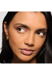 Primer plano del rostro de una mujer con piel lisa y bronceada, ojos marrones y cabello oscuro. Lleva un pequeño pendiente de aro dorado y tiene labios brillantes.