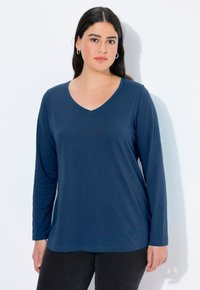 Kobieta z długimi ciemnymi włosami, nosząca srebrne kolczyki w kształcie kół, niebieską bluzkę z długim rękawem w serek i czarne spodnie na białym tle.