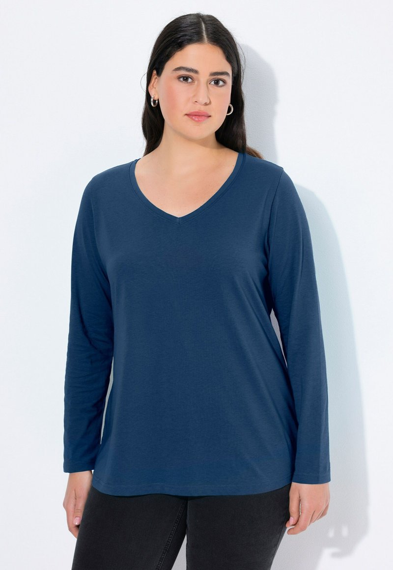 Kobieta z długimi ciemnymi włosami, nosząca srebrne kolczyki w kształcie kół, niebieską bluzkę z długim rękawem w serek i czarne spodnie na białym tle.