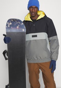 Rompevientos negro y gris con un forro amarillo, combinado con pantalones marrones. Sosteniendo una tabla de snowboard negra con detalles azules y guantes. Gorro azul.