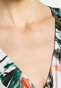 Blommönstrad topp med gröna och orange blad på en ljus bakgrund; design med V-ringning, synliga sömmar och mjuk tygstruktur.