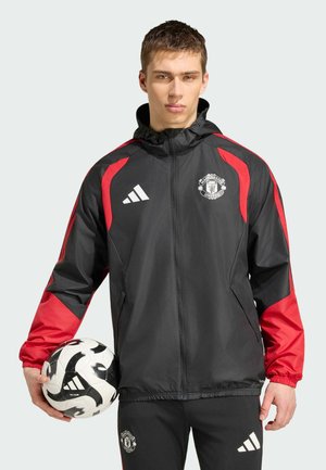 Młody mężczyzna w czarno-czerwonej kurtce Manchester United trzyma czarno-białą piłkę nożną Adidas.