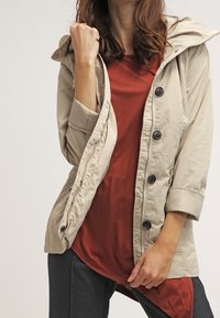 Chaqueta con capucha beige de diseño ligero, con cierres de botón y una textura suave, llevada sobre una blusa color óxido y pantalones oscuros.