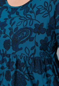Haut en tissu teal présentant des motifs floraux et paisley bleu foncé, avec une taille froncée et un col rond. Texture douce.