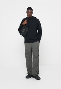 Felpa nera con cappuccio e cordini, pantaloni cargo grigi con più tasche e scarpe nere. Il modello tiene una borsa a tracolla nera.